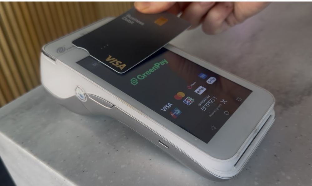 A hand tapping a Visa business debit card on a white GreenPay EFTPOS terminal